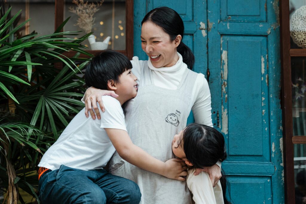 Happy cheerful mother bonding with cute son and daughter in casual wear while standing together against old shabby blue door and green garden plants