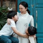 Happy cheerful mother bonding with cute son and daughter in casual wear while standing together against old shabby blue door and green garden plants